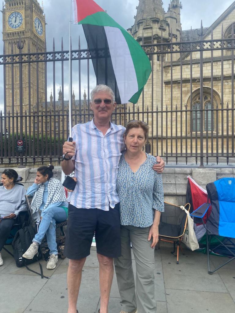 A man and a woman pose in front of the Houses of Parliament, holding a Palestinian flag. Other participants can be seen in the background.