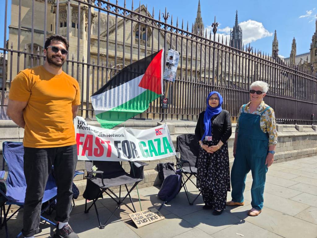 Three individuals standing in front of a banner that says 'FAST FOR GAZA,' with a Palestinian flag waving in the background. The setting appears to be outside a historic building.