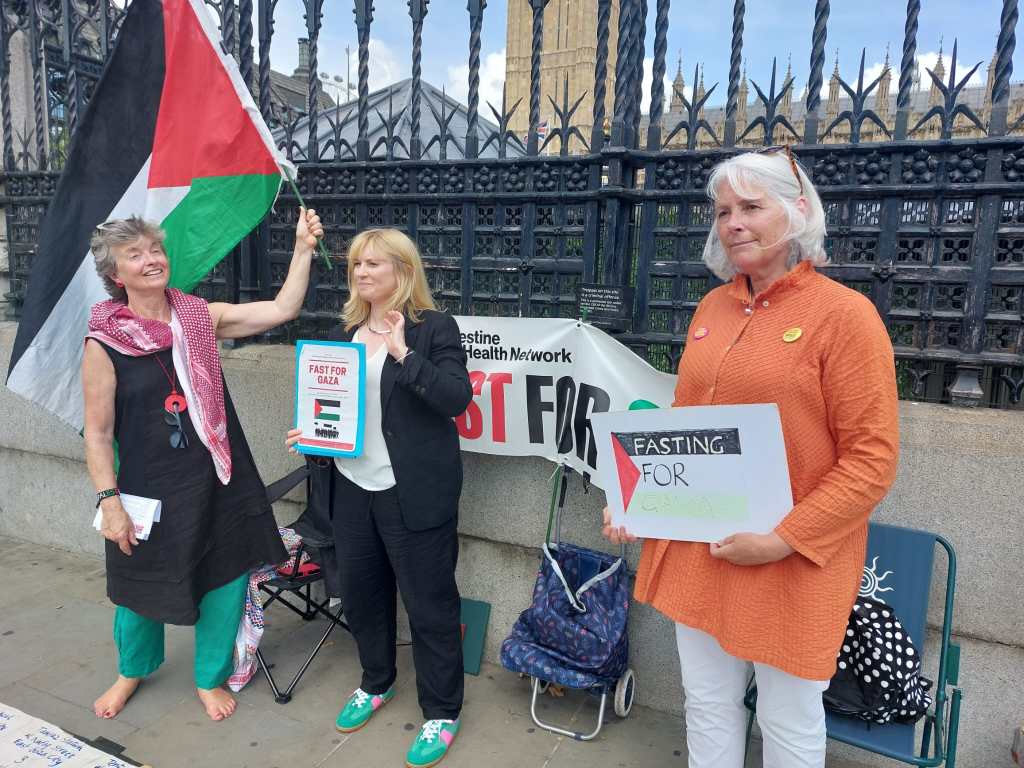 Three women participating in the Fast for Gaza demonstration, holding signs and a Palestinian flag outside Parliament.