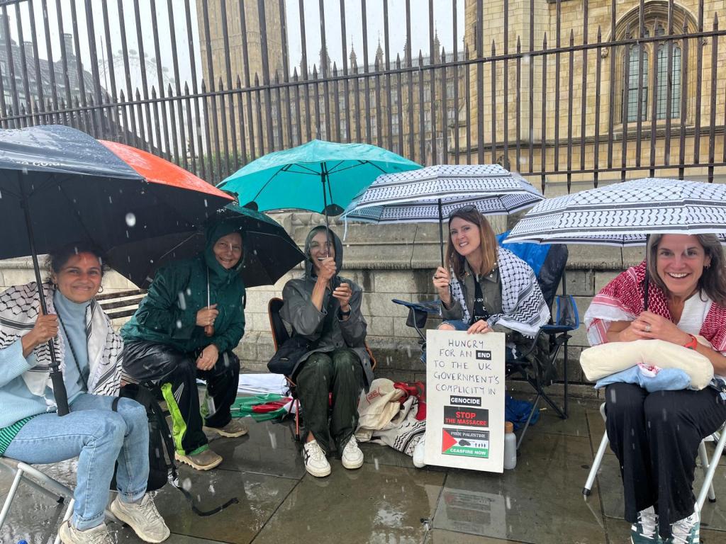 A group of five individuals sitting outside Parliament under umbrellas, holding a sign that reads 'HUNGRY FOR AN END TO THE UK GOVERNMENT'S COMPLICITY IN GENOCIDE'. They are dressed in raincoats and display solidarity with Palestine.