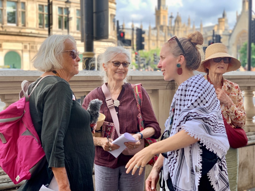 A group of women engaged in conversation outdoors, with one wearing a traditional scarf and another carrying a pink backpack, set against a backdrop of Parliament buildings.
