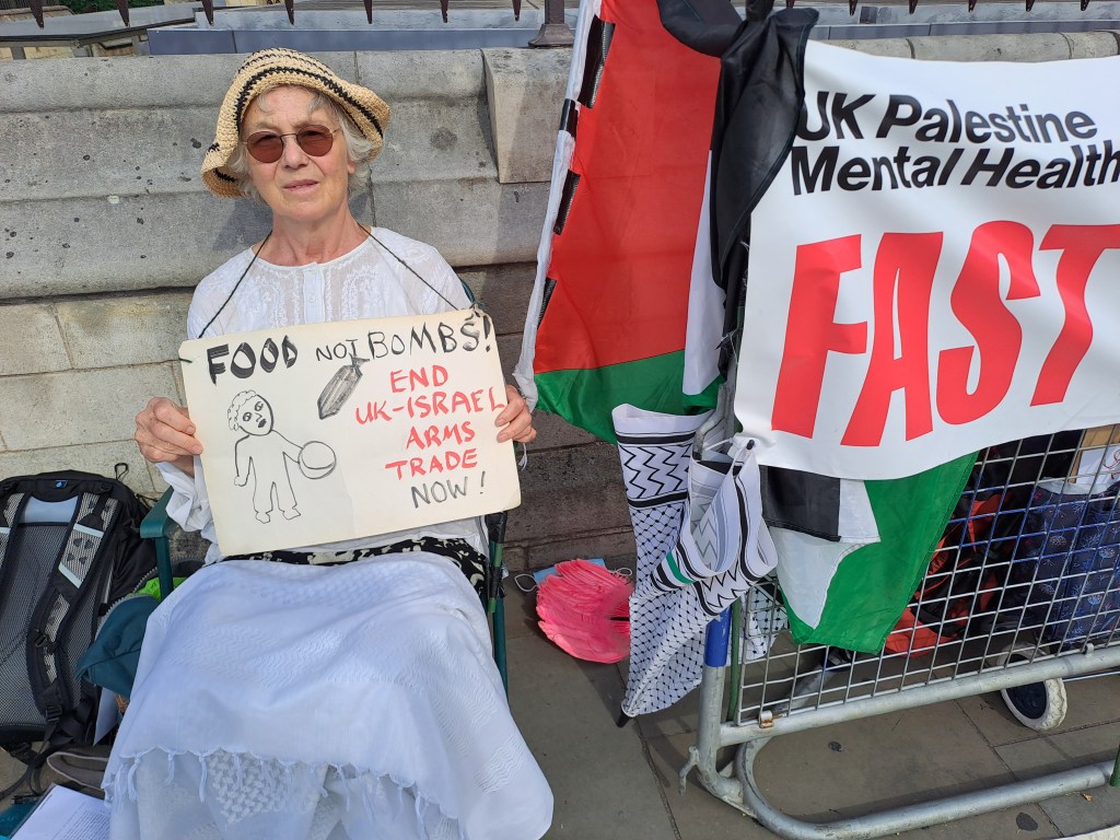 An elderly woman sitting on a chair holding a sign that reads 'FOOD NOT BOMBS! END UK-ISRAEL ARMS TRADE NOW!' She is wearing glasses and a hat, surrounded by flags related to the Palestinian cause.