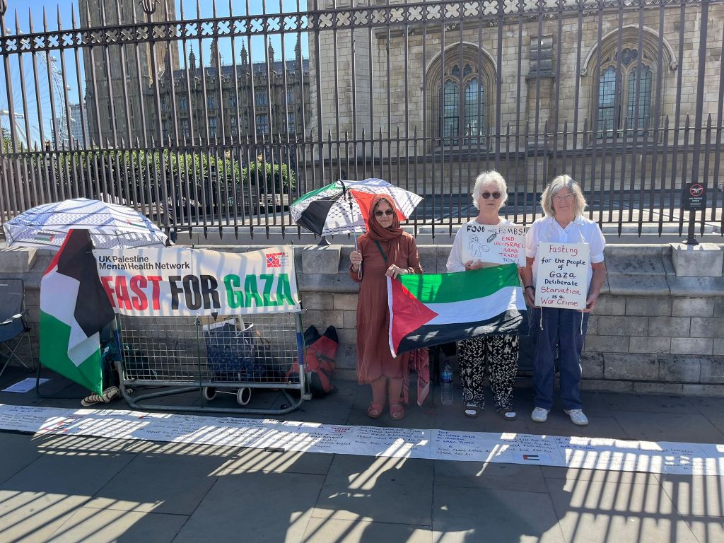 Four individuals participating in the Fast for Gaza event, holding a Palestinian flag and holding signs, in front of the Parliament building.